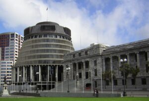 A photograph of the Beehive and Parliament in Wellington. The beehive is on the left and parliament on the right. The sky above has puffy white clouds and blue peaking through.