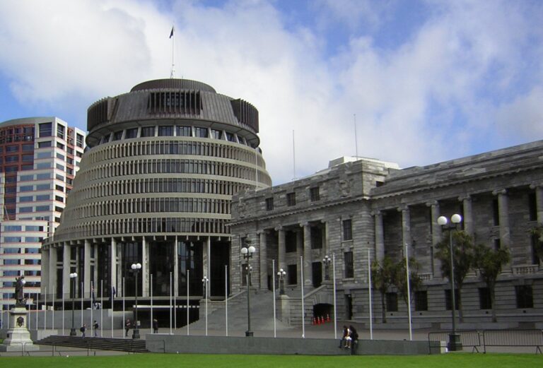 A photograph of the Beehive and Parliament in Wellington. The beehive is on the left and parliament on the right. The sky above has puffy white clouds and blue peaking through.