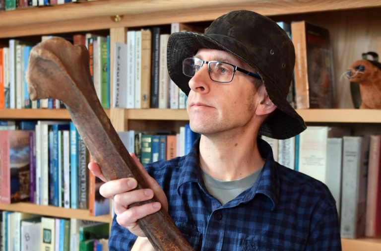 A photo of a man holding a large Moa bone. He is wearing a bucket hat and a blue and black checked shirt. He is looking up and to the left at the top of the moa bone. Behind him is a bookcase full of books.