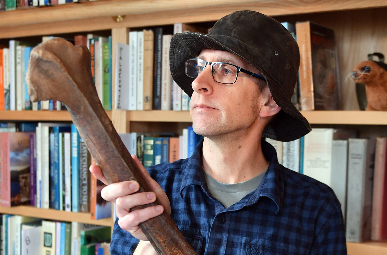 A photo of a man holding a large Moa bone. He is wearing a bucket hat and a blue and black checked shirt. He is looking up and to the left at the top of the moa bone. Behind him is a bookcase full of books.